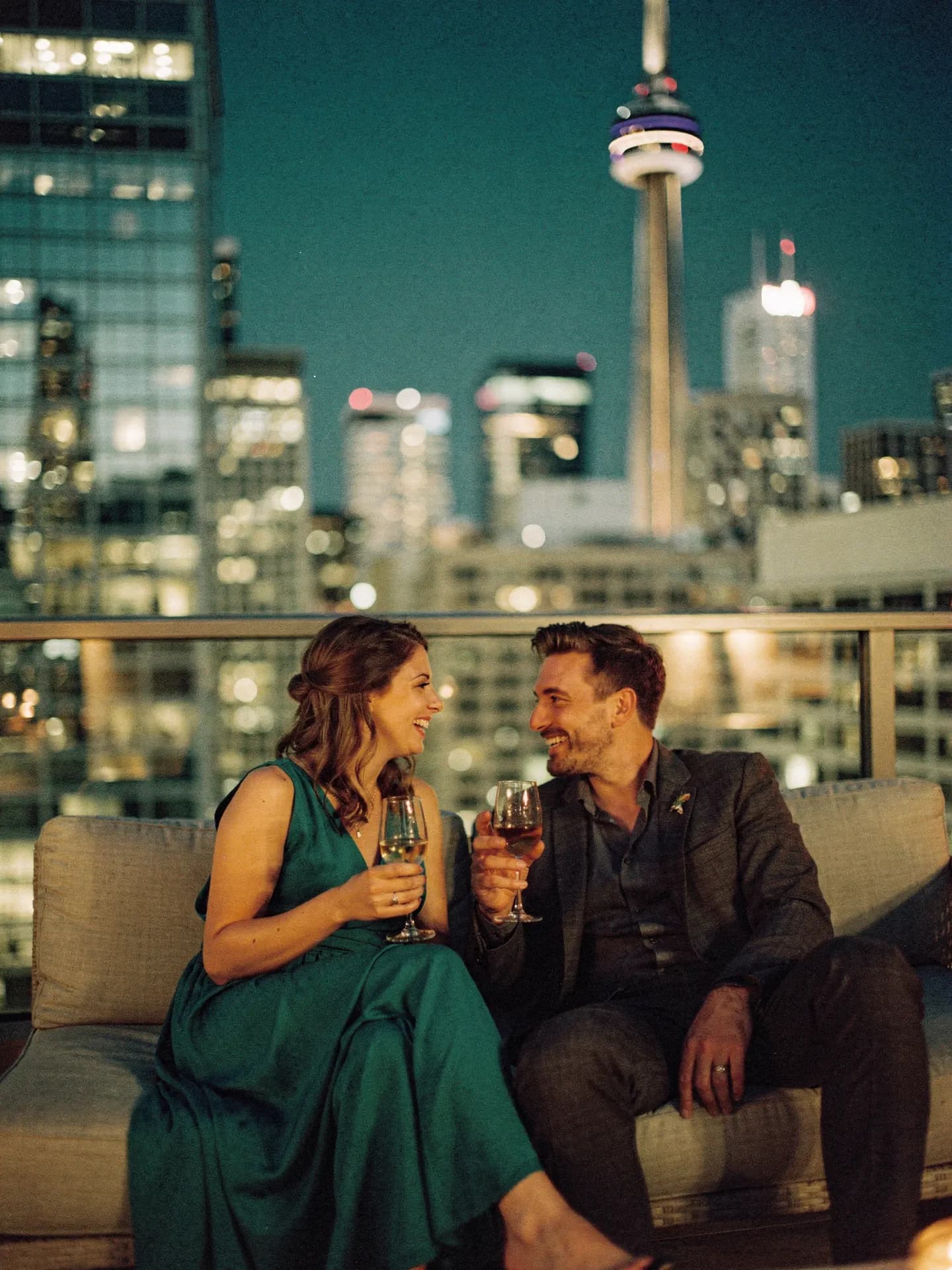 A couple with wine on a balcony at night, the Toronto skyline lit up behind them